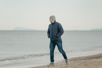 A muscular man in the grey hat, jeans, bologna jacket, and sneakers walks near the Mediterranean Sea