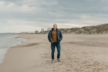 A muscular bald stylish man in the down jacket, jeans and sneakers walks with hands in jeans pockets on the seashore in the afternoon