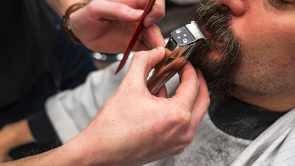Bearded man in barbershop. Haircut concept. Man visiting hairstylist in barbershop. Barber works with hair clipper. Hands of barber with hair clipper, close up.