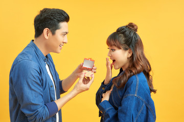 love, couple, proposal and people concept - man giving diamond engagement ring in little red box to happy woman over  yellow background