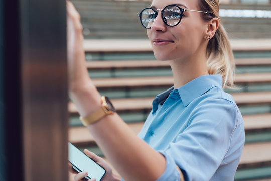 Interactive digital kiosk with public transport Info. Smiling female standing at big display with smartphone in hand.Young woman touching with finger interactive futuristic screen. Advanced technology