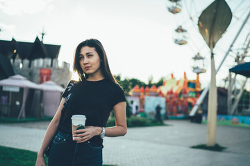 Thoughtful asian hipster girl in casual wear feeling tired after visiting amusement park standing with coffee to go cup on street, female traveler sad about vacation ending leaving fun fair .