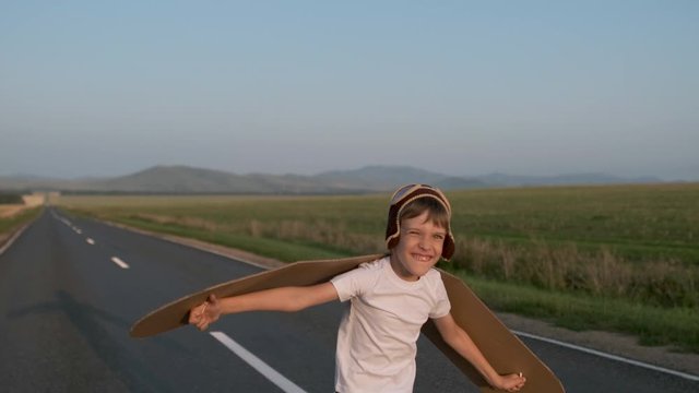 Happy smiling little boy child aviator pilot of airplane running with toy cardboard plane wings in road. Kid dreams of flying, traveling in summer nature at sunset outdoors, travel dream, 4 K slow-mo