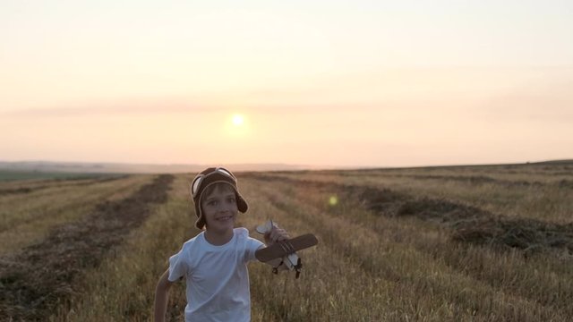 Happy little boy child in helmet aviator pilot of airplane running with toy wooden plane in field. Kid dreams of flying, traveling in summer nature at sunset outdoors. travel freedom dream 4 K slow-mo