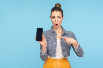Portrait of funny amazed fashionably dressed woman with hair bun pointing at mobile phone and...