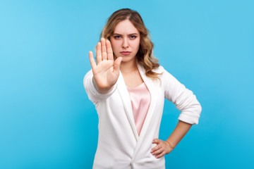 Prohibition concept. Portrait of serious woman with wavy hair in white jacket doing stop gesture, warning sign with raised palm, enough, showing limit. indoor studio shot isolated on blue background