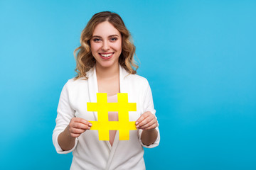 Hashtag and blogging. Portrait of amazing beautiful positive woman with wavy hair in white jacket holding big paper hash symbol and smiling at camera. indoor studio shot isolated on blue background