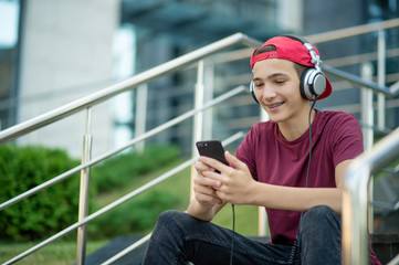 Young man listen to the music in a big earphones, in the street.  Happy teenage boy with mobile phone and headphones, outdoors. Cheerful teenager in casual clothes with cell phone.