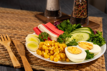Salad plate on the table with wooden cutlery. Crab sticks, boiled egg, corn and greens.