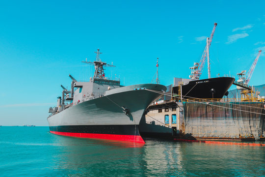 The military ship was waiting to be repair moored alongside in shipyard near floating dock on blue sky.