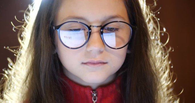 Close up portrait of smart schoolgirl with long brown hair wearing  glasses. Girl surfing internet. Laptop screen reflect  in glasses. Concept of eye fatigue and ophthalmic problems. Slow motion