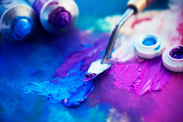Tubes of blue and purple oil paint lie on a palette, along with a thin palette knife that mixes the...