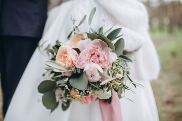bride in white dress holds in her hands bouquet of peonies, roses and greenery with pink ribbon