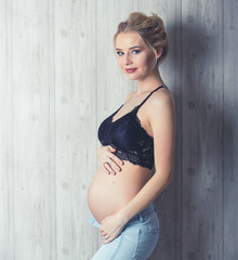 Luxurious pregnant blonde in jeans and black bustier posing against a wooden wall