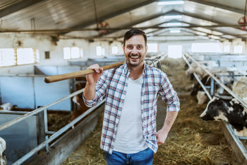 Handsome smiling unshaven caucasian farmer standing in stable with hay fork on shoulder. In...