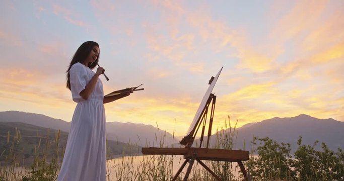 Professional female artist is inspierd by beautiful mountain lake in sunset, creating a painting on easel, enjoying her hobby - recreational pursuit, inspirational landscape concept 4k footage
