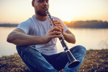 Young handsome bearded caucasian blond man sitting on cliff and playing clarinet. In background is river and sunset. - obrazy, fototapety, plakaty