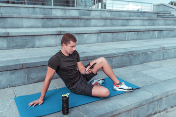 Fototapeta premium male athlete, in summer city, resting after workout, holding a smartphone, reading and writing message on smartphone, resting on a fitness mat. Bottle shaker with protein and water.