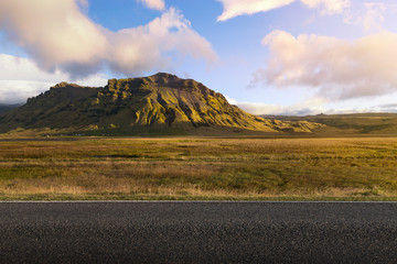 Sunset scene with background of big green mountain behind and empty road for foreground. Grass field in middle with warm tone of picture. Idea to put some product with beautiful landscape background
