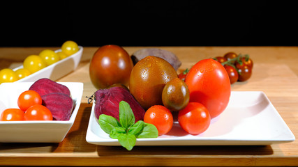 close up of variants of tomatoes and raw beetroot, covered with fresh water drops, on white tableware arranged on a wooden board, in front of a dark background