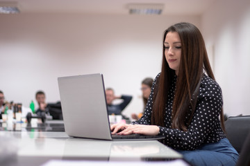 Young adult caucasian brunette woman sitting in an office environment looking at the laptop doing work. Multiple people in the background during a meeting.