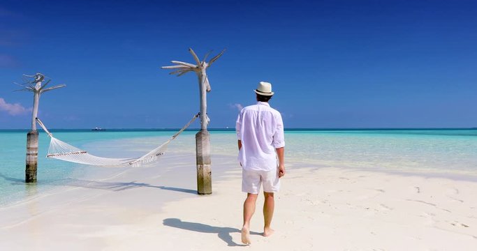 Ein Mann in wei&szlig;er Sommerkleidung l&auml;uft auf einem tropischen Traumstrand mit t&uuml;rkisem Meer und H&auml;ngematte und geni&szlig;et den Ausblick auf den Horizont