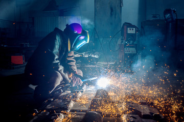 Side view of welder in protective helmet welding metal detail with sparks at factory  