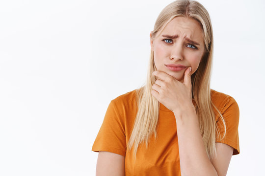 Skeptical displeased young blond girl, furrowed eyebrows and squinting, touch chin and look camera bothered and distressed, hear bad news, negative results, disappointing over white background