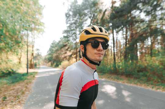 Closeup Portrait Of Beautiful Cyclist In Sunglasses And Helmet Standing Against Forest Road Background. Man In Sportswear Stands In The Park And Looks Away.