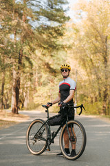 Obraz premium Handsome man in a bicycle suit stands with a bicycle on the road in a beautiful forest and poses at the camera, wearing a helmet and sunglasses. Portrait of a cyclist in the forest