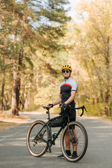 Handsome man in a bicycle suit stands with a bicycle on the road in a beautiful forest and poses at the camera, wearing a helmet and sunglasses. Portrait of a cyclist in the forest