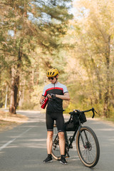Cyclist stands with a bicycle on the road in the forest and opens a bottle of water. Thirsty man rides his bicycle through the autumn forest and begins to rest and drink water