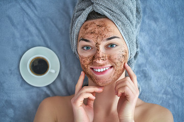 Portrait of smiling healthy woman in bath towel with natural cleansing face coffee scrub during spa day and skin care routine at home