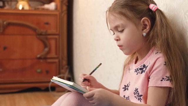 Little girl sit on floor, painting or writing. Serious preschooler with pigtails do homework. Dressed in pink t-shirt and tights. Concept of school, self development, painting. Сupboard on background