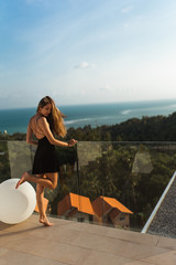 A beautiful girl in a dress is resting in a luxury villa by the pool with wine and in a hat taking cover from the sun. View of palm trees and the sea. Thailand, Koh Samui