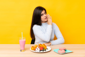 Young woman eating waffles and milkshake in a table over isolated yellow background whispering something