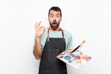 Young artist man holding a palette over isolated background surprised and showing ok sign