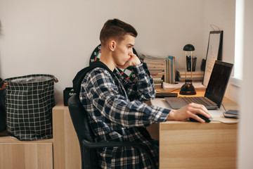 Teenage boy doing homework using computer sitting by desk in room alone