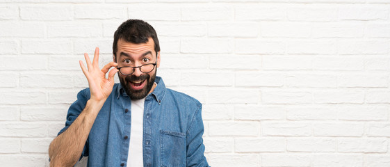 Handsome man with beard over white brick wall with glasses and surprised