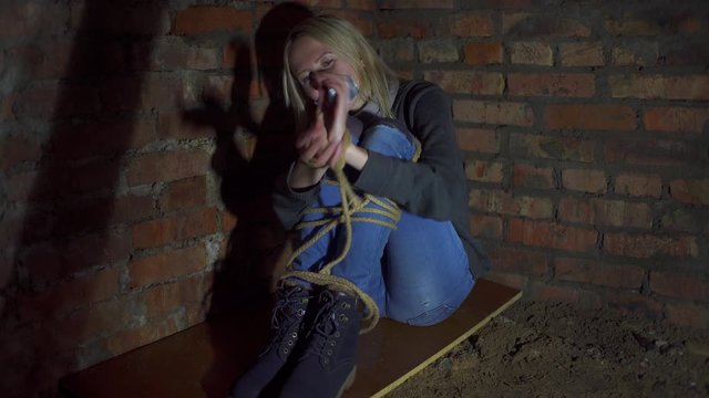 A kidnapped young blond woman tied is sitting in a basement on a concrete f...