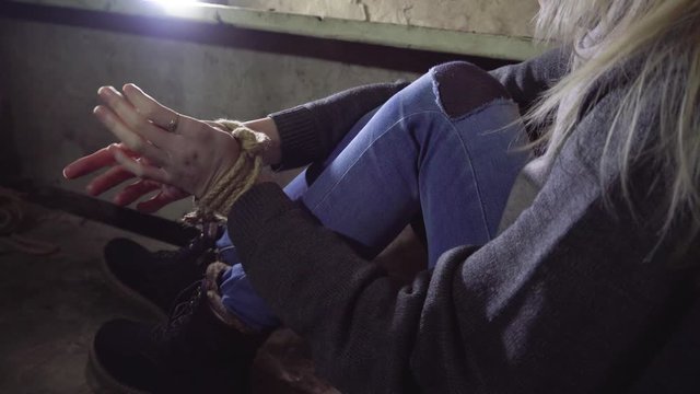 A kidnapped young blond woman tied is sitting in a basement on a concrete floor. Hands and feet tied with a rope. She's trying to free herself.