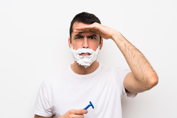 Man shaving his beard over isolated white background looking far away with hand to look something