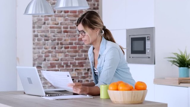Video of pretty young woman working with laptop and documents in the kitchen at home.