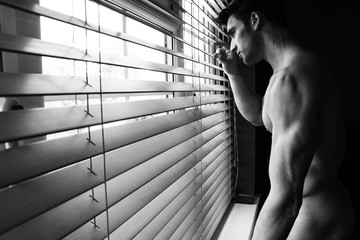 Portrait of naked handsome man with beard and muscular arms looking through blinds of window