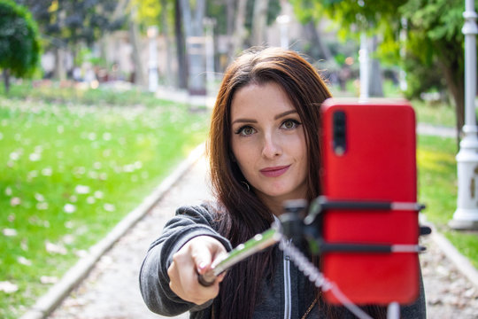Pretty attractive brunette with amber orange eyes in a public park taking a selfie with a selfie stick and a red smartphone looking at the camera