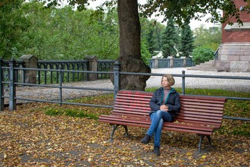 A young, pretty woman sits in a park on a bench, cross-legged, holding a telephone in her hand. Around are lying on the ground autumn fallen yellow leaves.