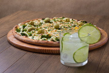 Broccoli pizza served on wooden board. Baked with mozzarella, broccoli, corn, peas and olives. Glass with carbonated water and lemon. Vegetarian. Horizontal photo with bottom alignment of the cup.