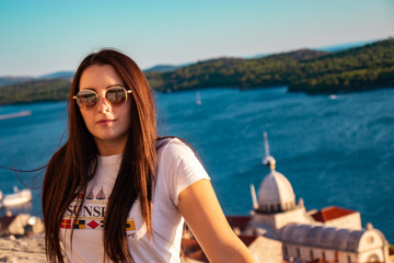 Fototapeta na wymiar Portrait of an attractive brunette with a white shirt and sunglasses. Leaning on a wall , adriatic sea and the SIbenik cathedral in the distance