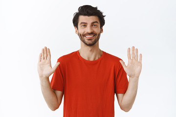 Fototapeta premium Relax, everything under control. Confident good-looking bearded guy in red t-shirt, raising hands up and smiling, asking keep calm, rejecting something politely, standing white background