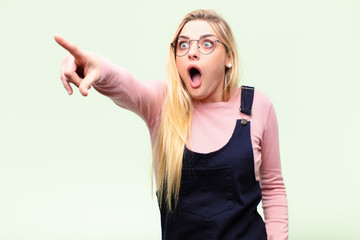 young pretty blonde woman feeling shocked and surprised, pointing and looking upwards in awe with amazed, open-mouthed look against flat wall